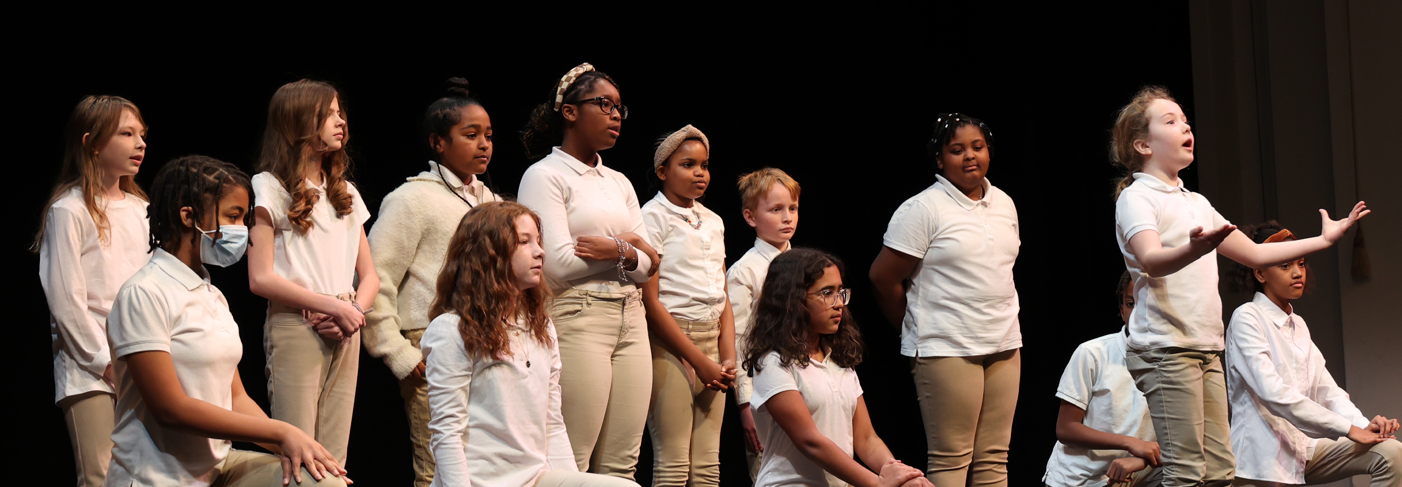 A group of schoolchildren in white shirts and tan pants stand or kneel on a stage.