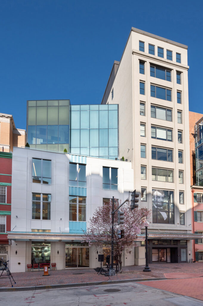 Street view of two buildings along a city street, the one to the right tall and tan, the one to the left squat and white.